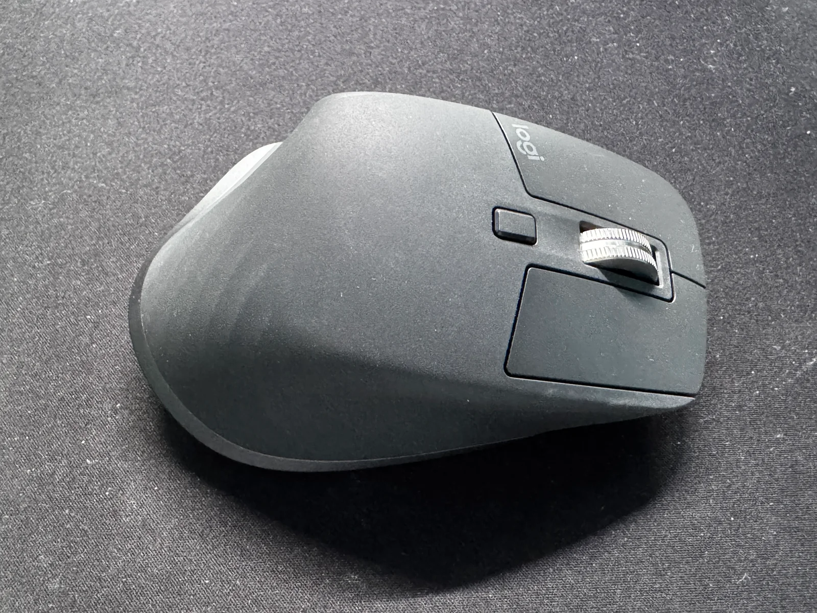 มุมด้านข้างของเมาส์ Logitech MX Master สีเทาเข้ม เห็นล้อเลื่อนนิ้วโป้งด้านข้างและปุ่มควบคุม มีคราบฝุ่นบนผิวสัมผัส