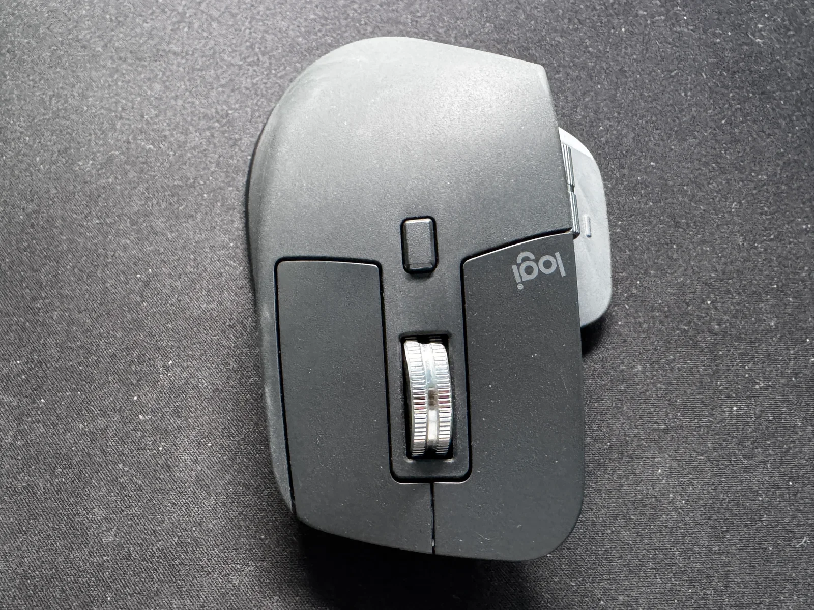 ด้านบนของเมาส์ Logitech MX Master สีเทาเข้มและเงิน แสดงปุ่มคลิกซ้ายขวา ล้อเลื่อนโลหะ และปุ่มปรับโหมด มีฝุ่นและรอยนิ้วมือ